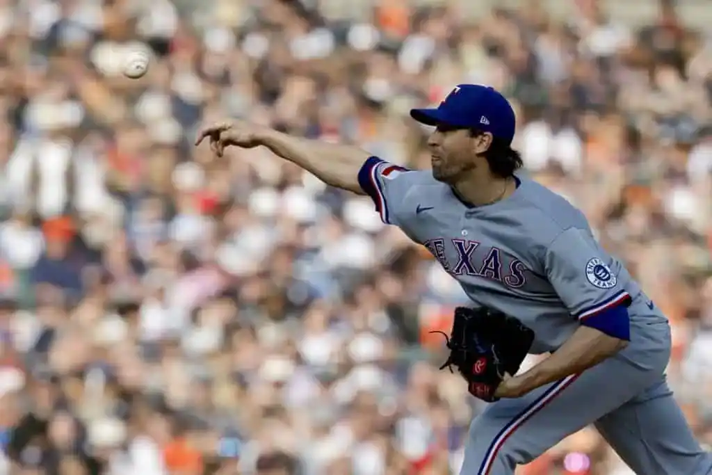 A Texas Rangers pitcher in a gray jersey throws a baseball toward home plate, with a crowd of fans in the blurred background.