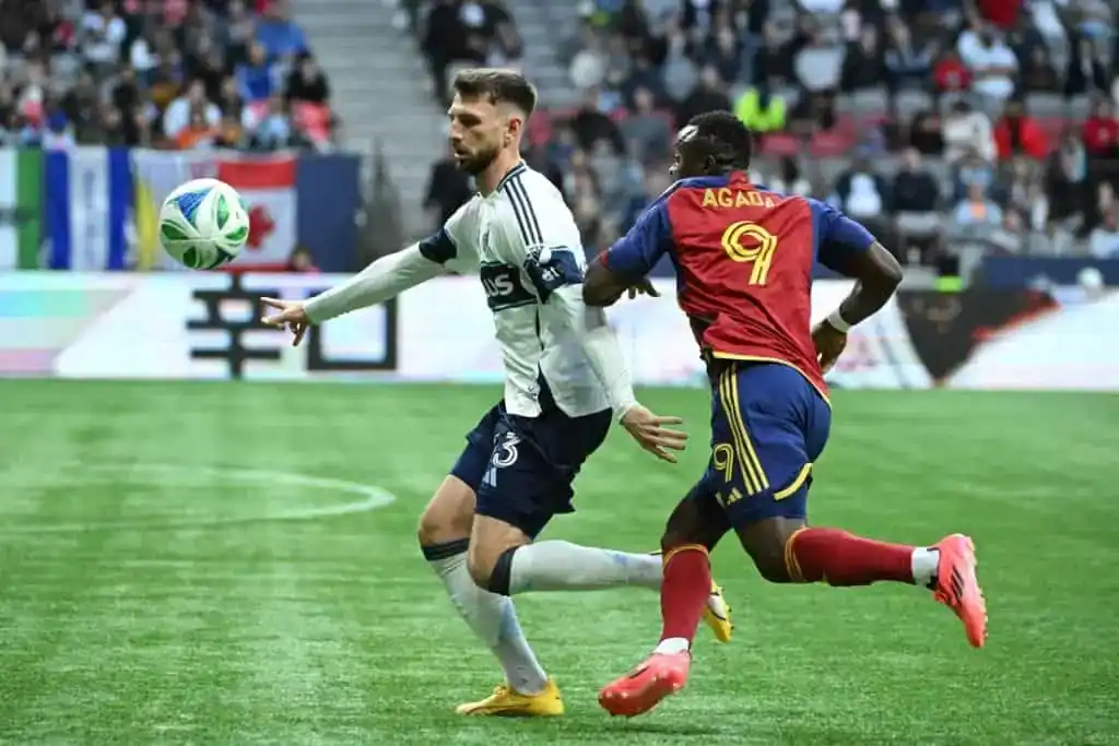 A soccer player in a white uniform passes the ball while being pursued by an opponent in a red and yellow jersey.