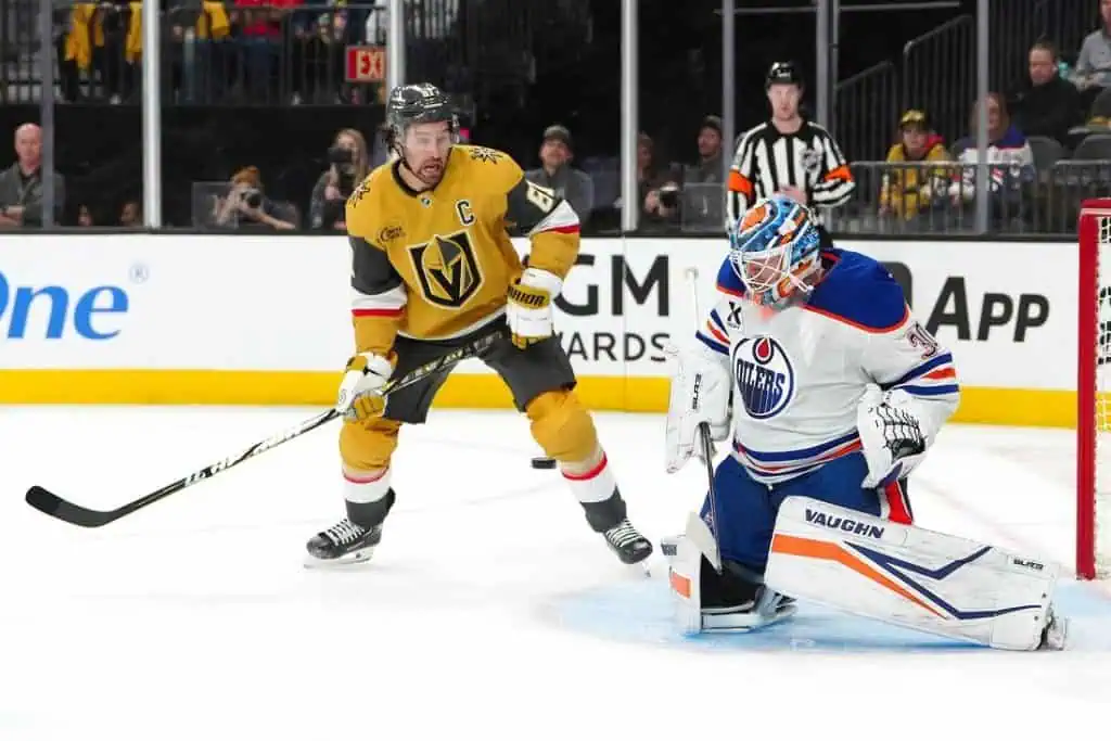 A hockey game scene with a player in a gold jersey preparing to take a shot, while the goalie in blue and orange braces for action.