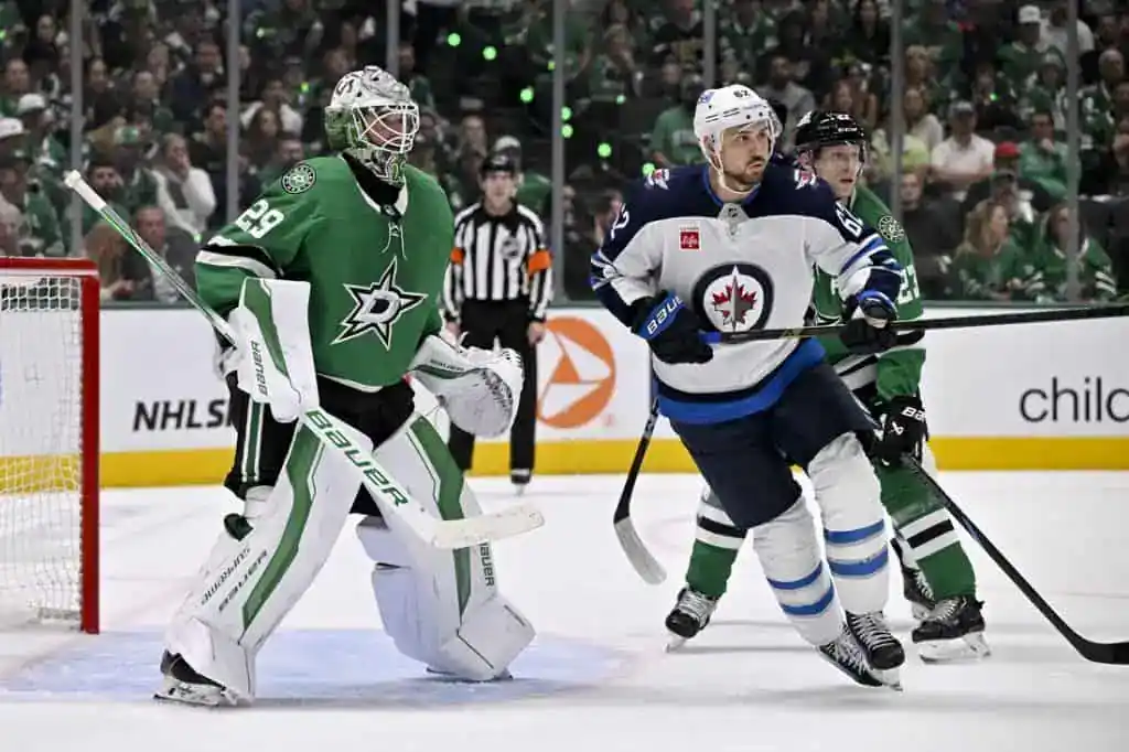 A tense hockey game scene featuring a goalie in green gear, a player in white with blue accents, and action taking place near the net.