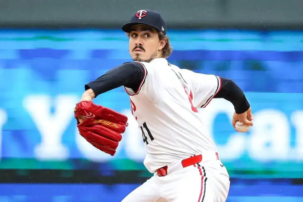A pitcher in a white baseball uniform prepares to throw a pitch, wearing a red glove and a cap with a logo, against a colorful backdrop.