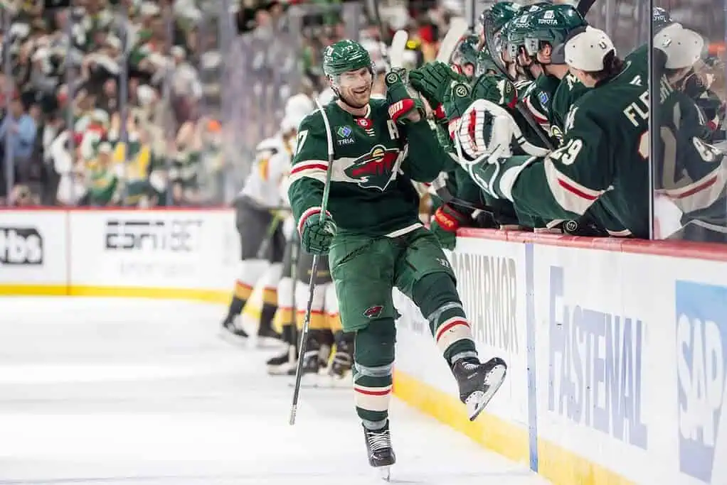 A Minnesota Wild player celebrates a goal, skating joyfully as teammates cheer enthusiastically from the bench in a packed arena.
