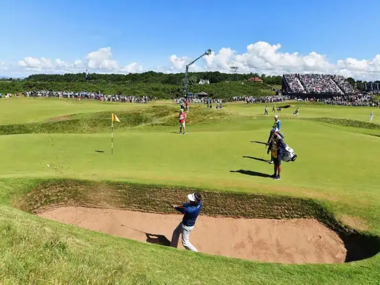 Golfista realiza un tiro desde un búnker durante el Open Championship 2024, con espectadores y cámaras siguiendo la acción en el campo.