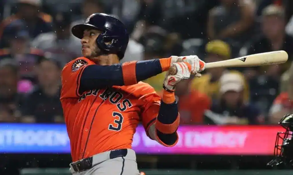 A baseball player in an orange jersey swings a bat during a game, with blurred fans visible in the background.