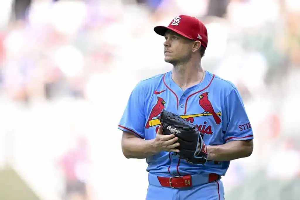 A baseball player in a light blue jersey with red accents stands on the field, holding a glove and preparing for play.