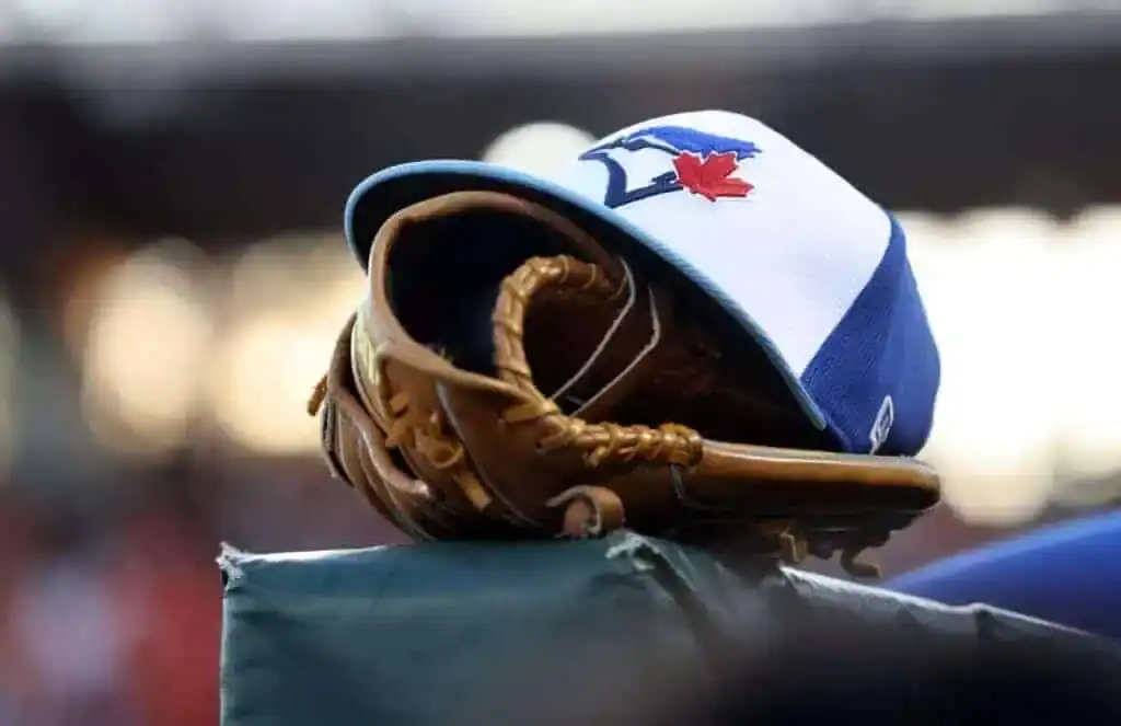 A blue and white baseball cap with the Toronto Blue Jays logo rests on a brown baseball glove, sitting atop a dugout edge.