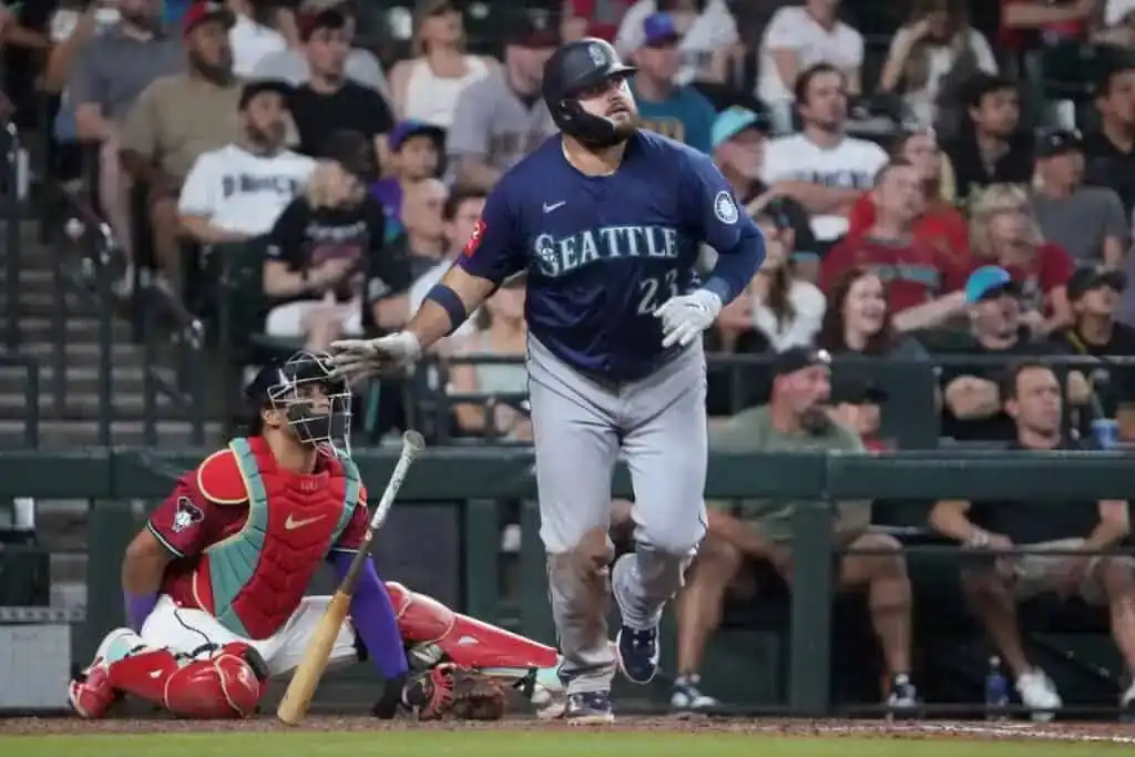 A Seattle baseball player runs toward first base, while the catcher crouches behind home plate during an intense game. Fans cheer in the stands.