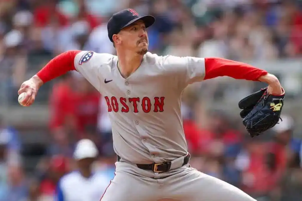 A Boston Red Sox pitcher in action, wearing a gray jersey with red sleeves, winding up to throw a baseball against a blurred crowd background.
