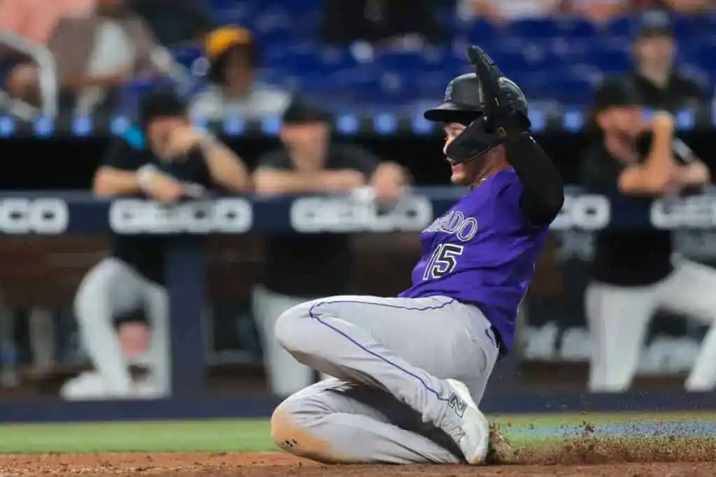 A player in a purple baseball uniform slides onto the base, with teammates in the background watching intently.