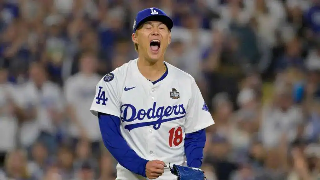 A Los Angeles Dodgers player in a white jersey with blue accents stands on the field, focused during a game night with a cheering crowd behind.