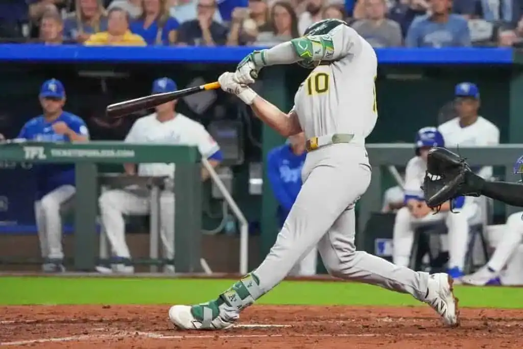 A baseball player swings a bat, making contact with the ball, while teammates and opponents watch from the dugout and field.
