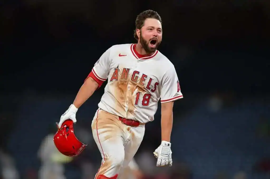 A player in a stained Los Angeles Angels uniform walks confidently on the field, holding a red helmet, amidst a blurred stadium backdrop.
