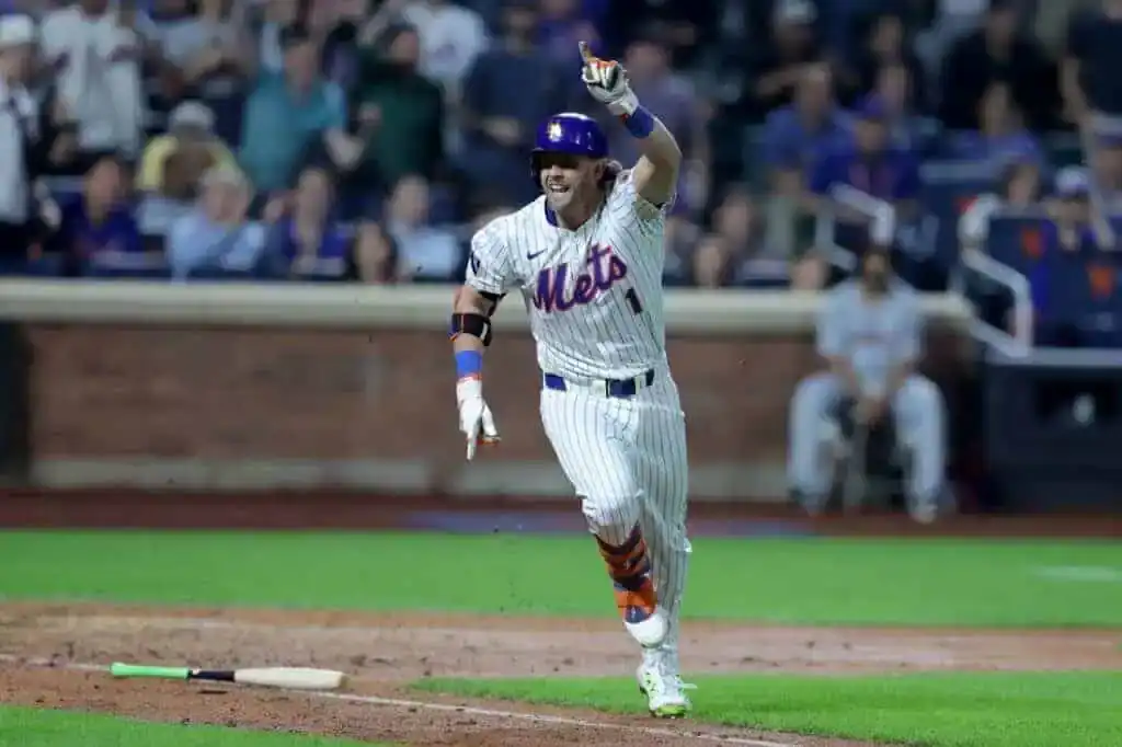 A New York Mets player celebrates and points towards the crowd while running on the baseball field, with fans cheering in the background.