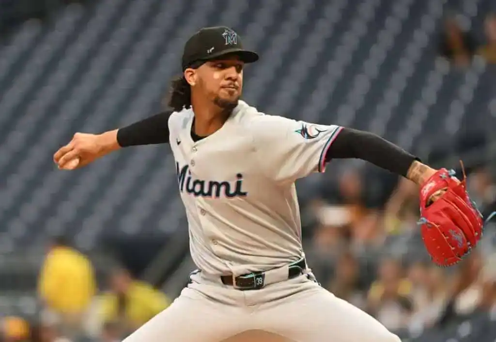 A Miami Marlins pitcher winds up to throw a baseball, showcasing his red glove against a blurred stadium crowd in the background.