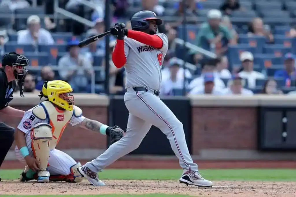 6 A baseball player in a gray Washington jersey swings at a pitch while a catcher in a yellow helmet crouches behind home plate.
