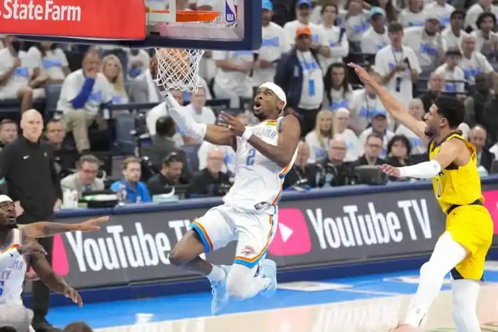 A basketball player in a white and blue jersey attempts a layup while being guarded by an opponent during a dynamic game scene.