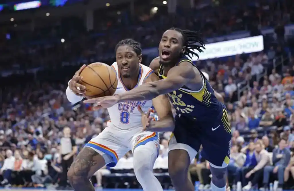 Two basketball players compete for the ball during a game, with one player in a white uniform holding the ball and the other in a dark uniform defending closely.