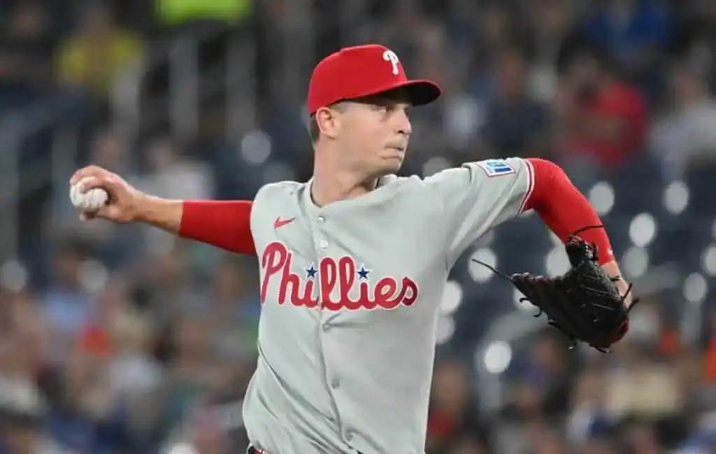 A Philadelphia Phillies player in a gray uniform winds up to pitch on a baseball field, with spectators blurred in the background.