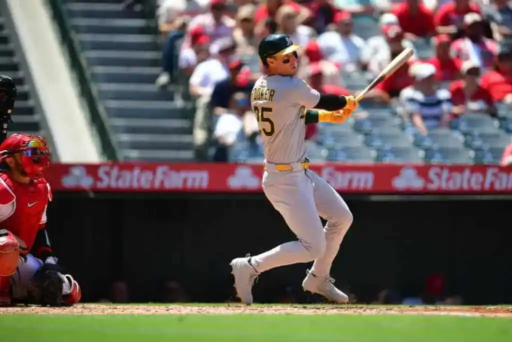 A baseball player in a grey uniform swings a bat, with a red catcher behind him and spectators in the stands.