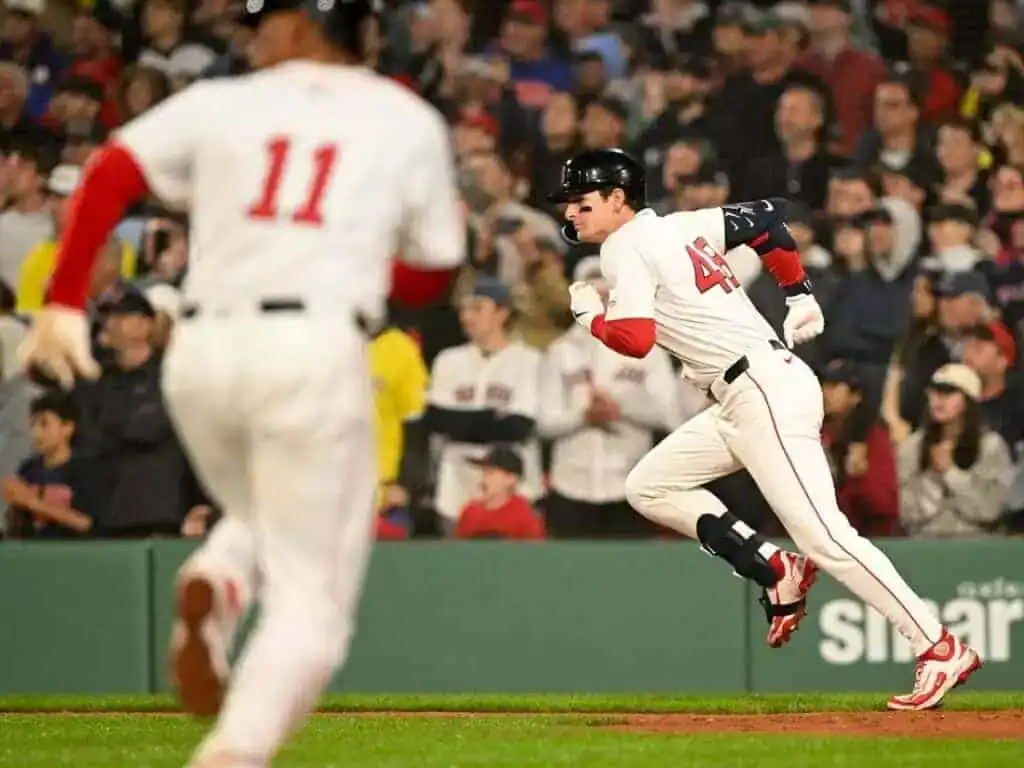 A Red Sox player in white uniform runs toward first base with fans cheering in the background at a baseball game.