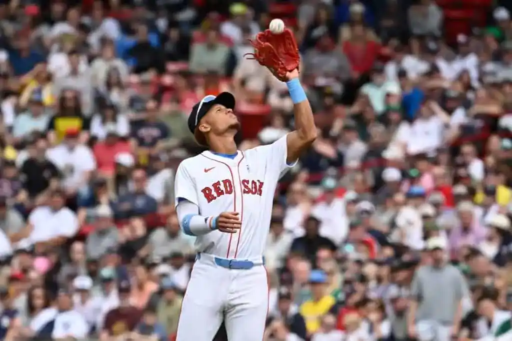 A Boston Red Sox player in a white uniform leaps to catch a baseball, with a packed crowd cheering in the background.