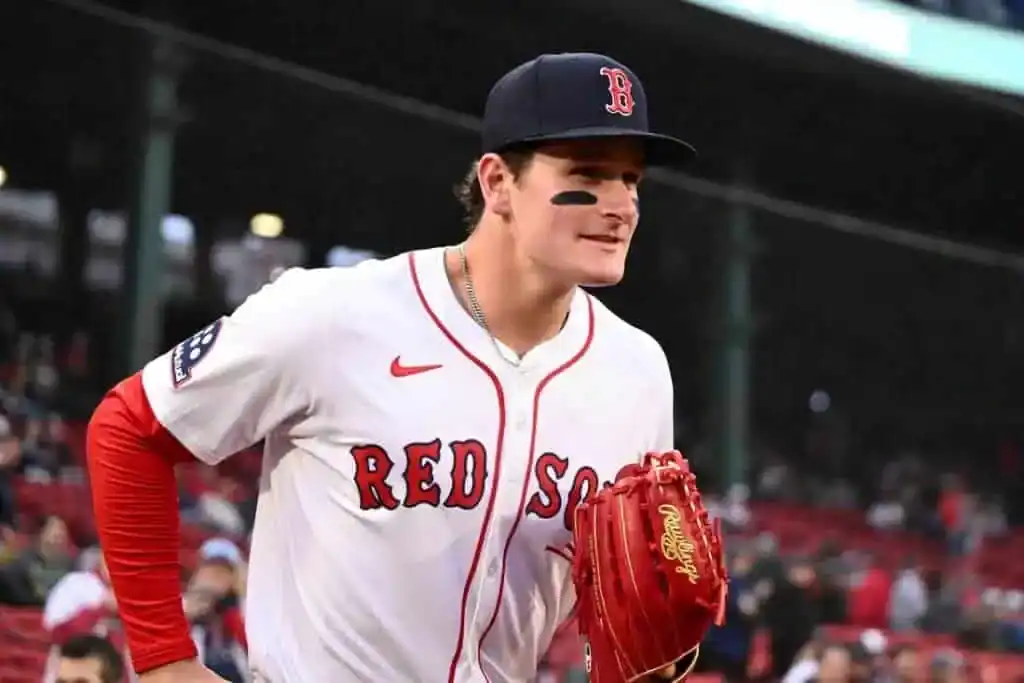 A baseball player in a red and white Boston Red Sox uniform walks onto the field, holding a glove, with Fenway Park in the background.