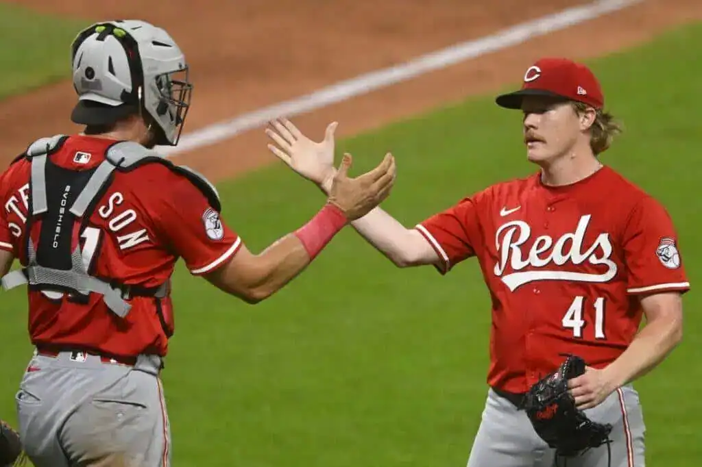 Cincinnati Reds players celebrate a successful play on the field, with one in a catcher's gear and the other ready to pitch.