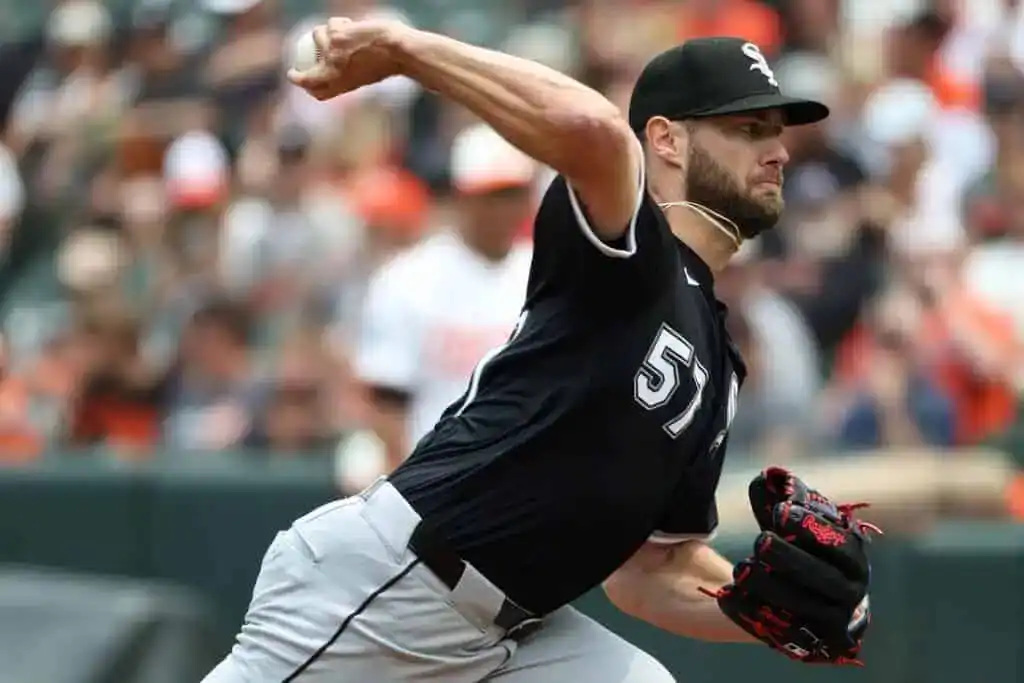 A pitcher in a black and white uniform delivers a fastball on the field, with fans cheering in the background.
