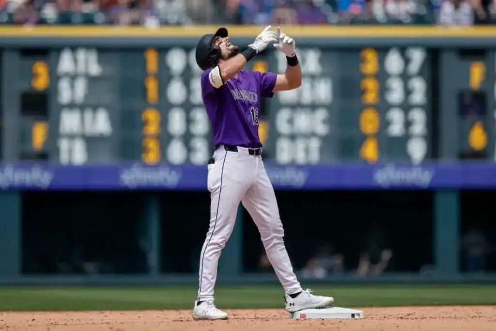 A baseball player in a purple jersey celebrates at first base, with a scoreboard displaying game scores in the background.