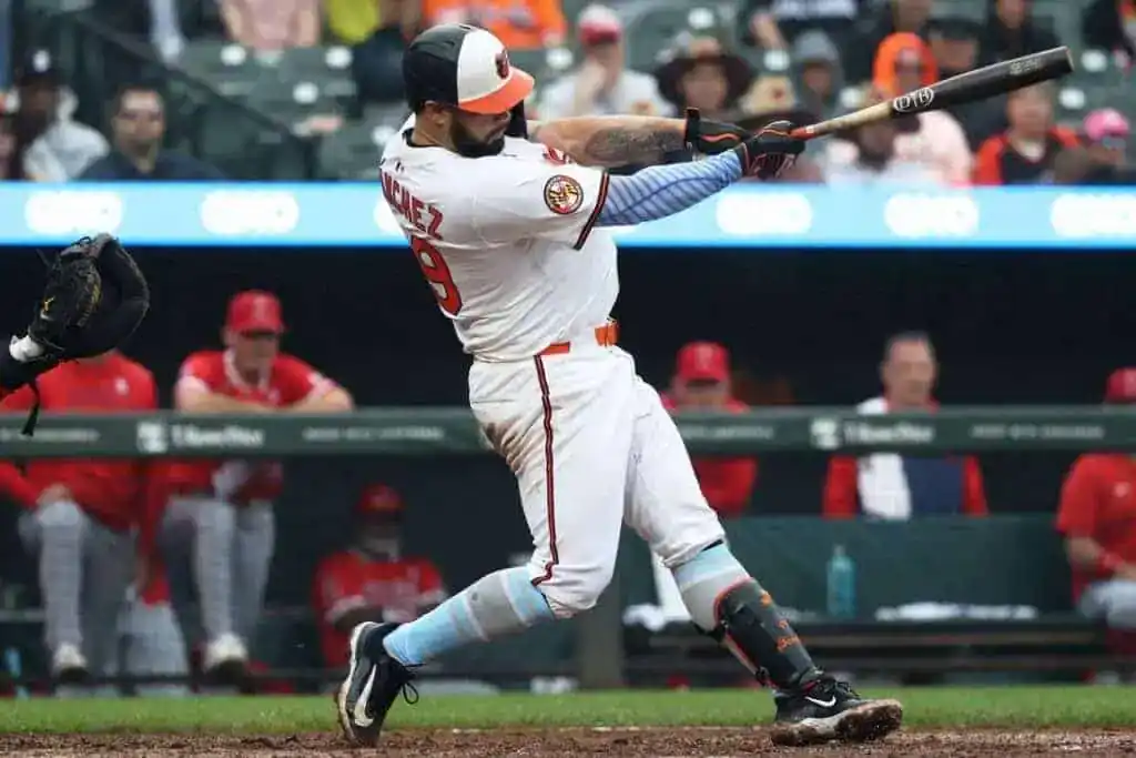 A baseball player in a white uniform swings a bat, preparing to hit a pitch, while teammates in red jerseys observe from the dugout.