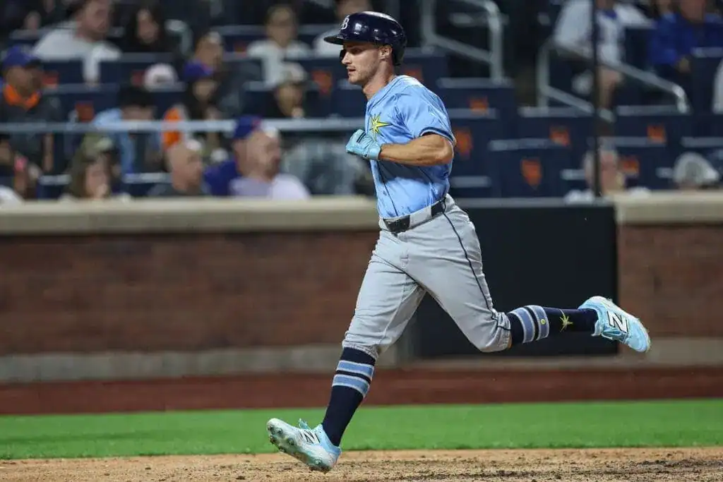 A baseball player in a light blue jersey and gray pants runs towards first base, with fans in the background.