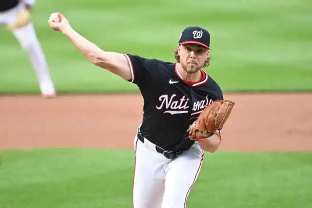 A baseball pitcher in a black Nationals jersey winds up to throw a pitch on a grassy baseball field.