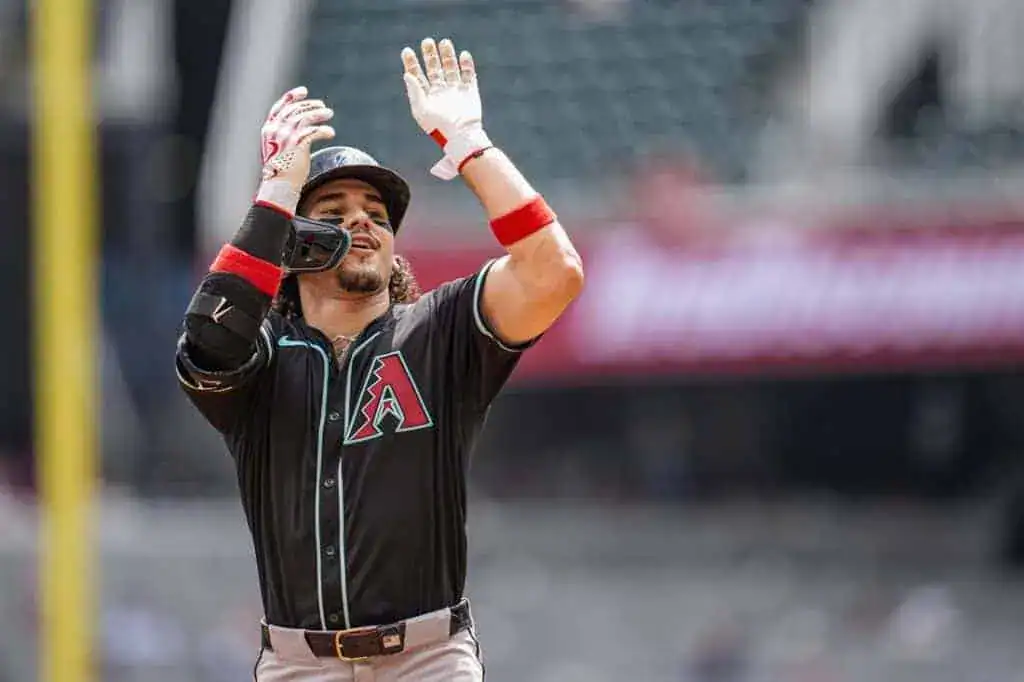 A baseball player in a black jersey with teal and red accents celebrates enthusiastically on the field, lifting both arms up.